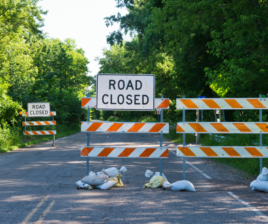 road closed sign attached to orange and white striped barricades on a street lined with trees